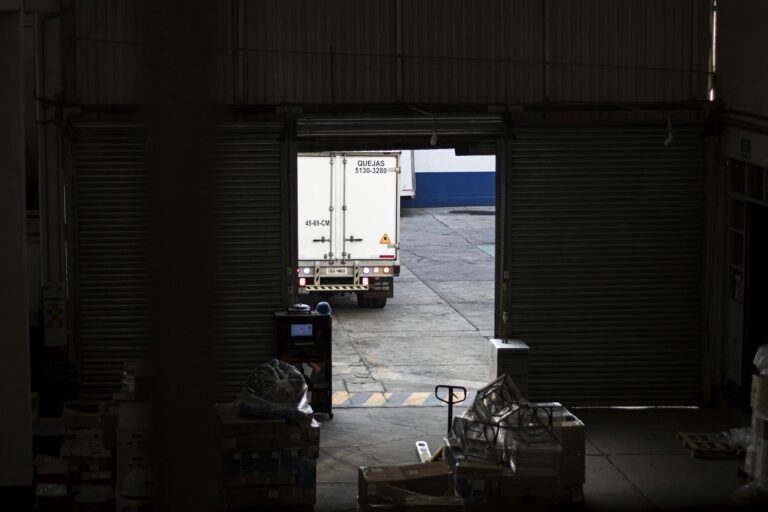 A white delivery truck exits a dimly lit warehouse loading dock, surrounded by packages and pallets.